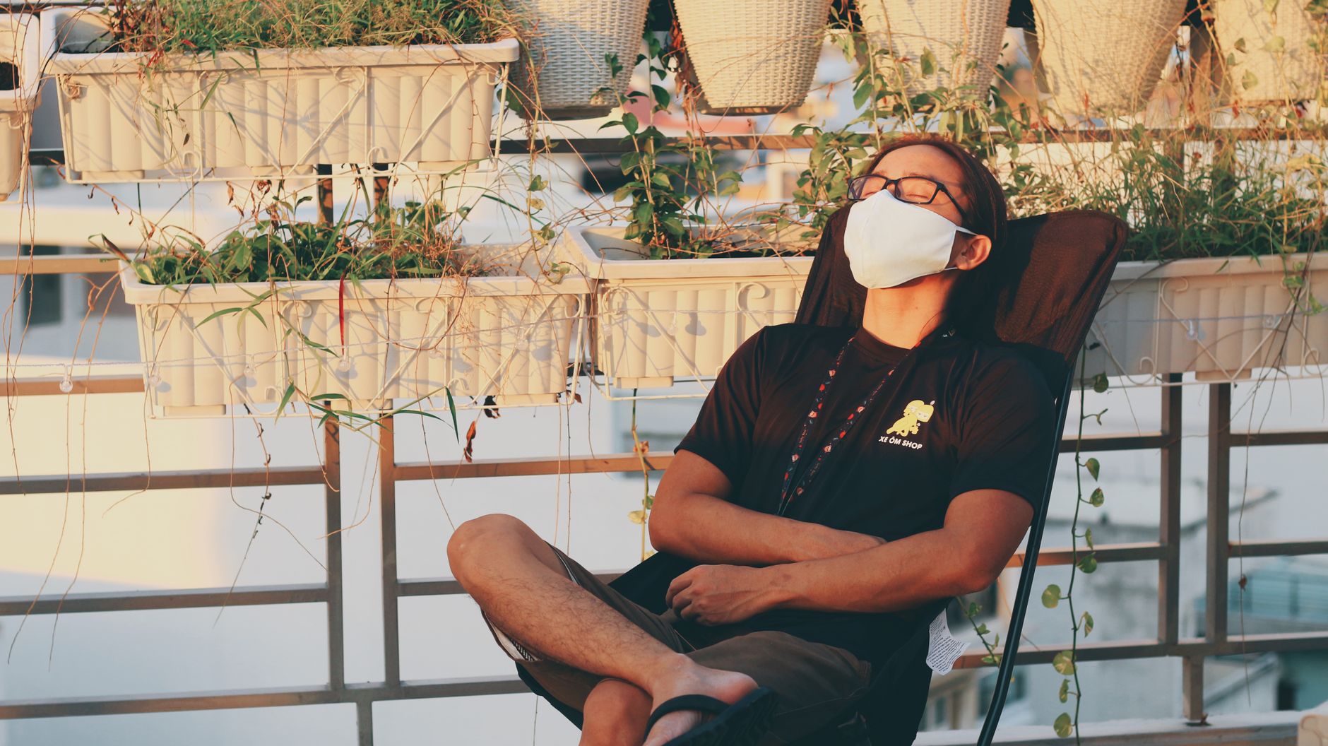 A man in a face mask relaxes on a sunny balcony with plants, embracing a moment of calm.
