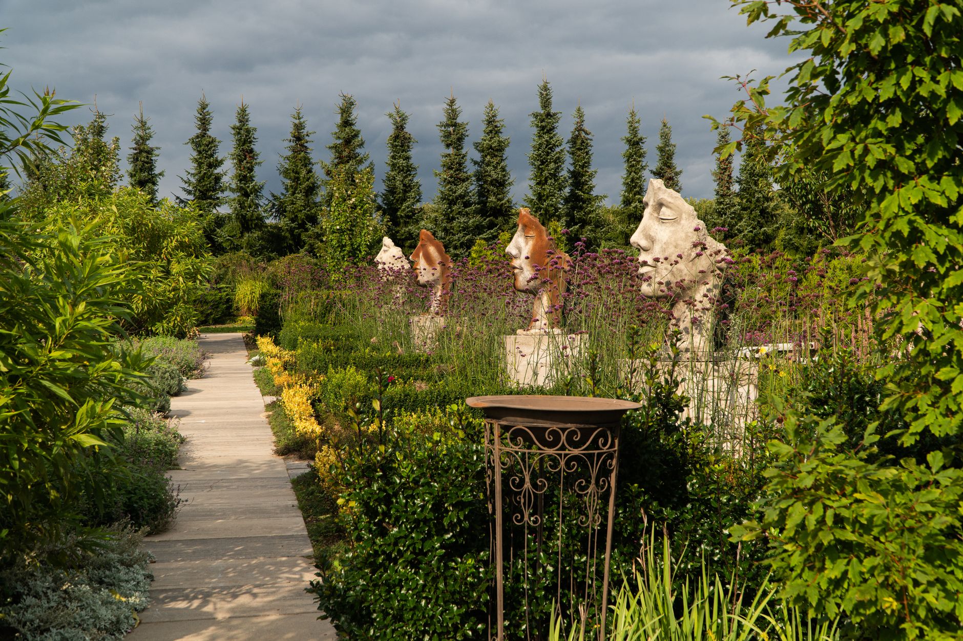 Explore a tranquil garden path featuring unique sculptures and lush greenery under a cloudy sky in Dobrzyca, Poland.