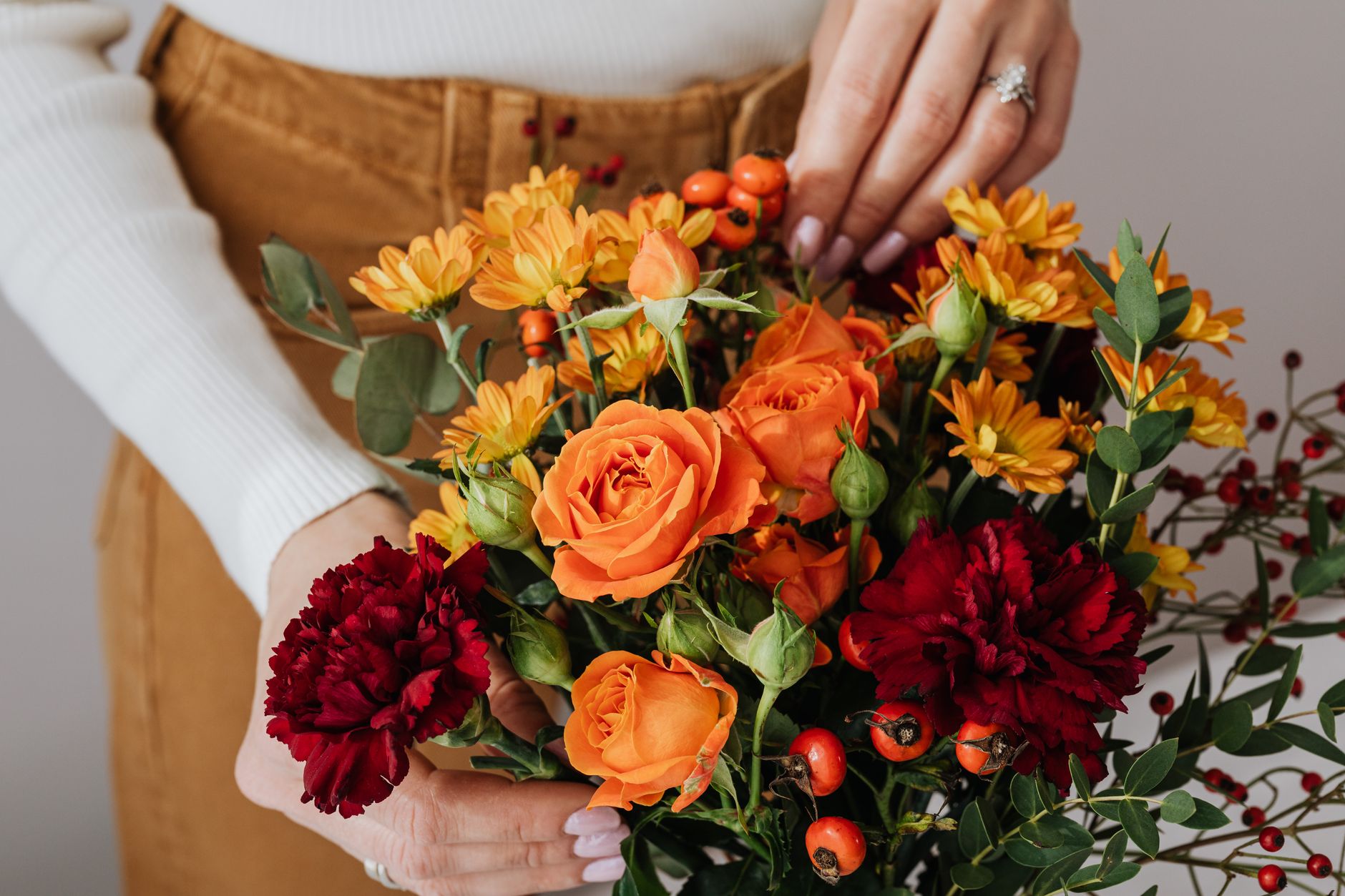 Beautiful autumn bouquet with orange roses, red carnations, and green foliage held by a woman's hands.